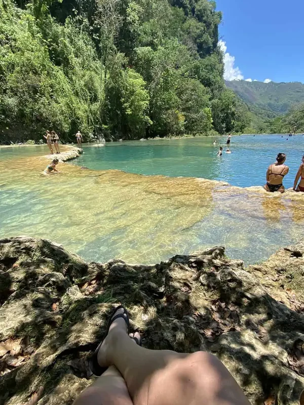 A first-person perspective showing a woman's legs as she sits on the rocky edge of a natural pool in Semuc Champey. The clear green waters stretch out in front of her, bordered by lush greenery and a vibrant forest scene, reflecting a peaceful and relaxing atmosphere.