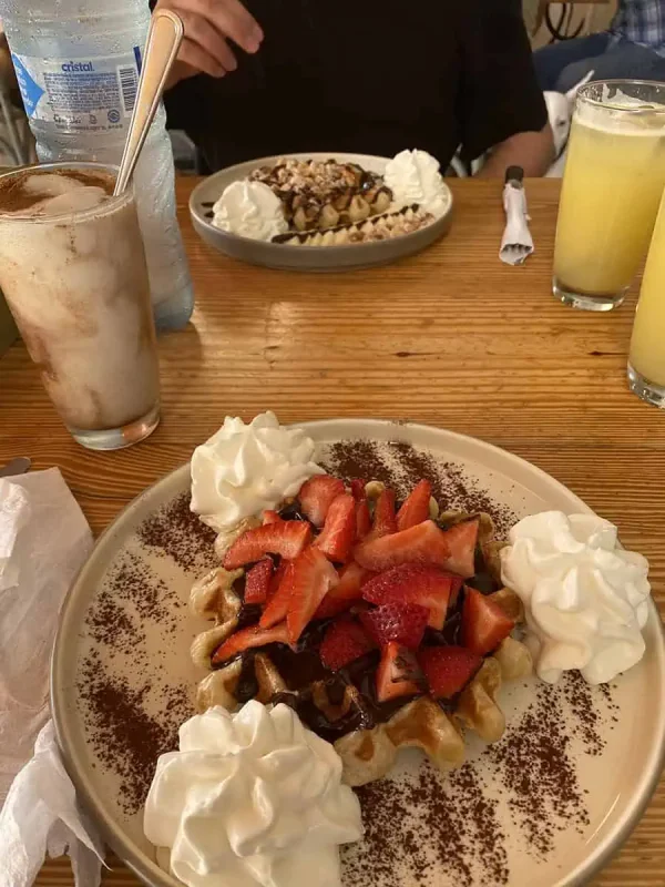 A mouth-watering display of desserts on a wooden table, featuring a waffle topped with fresh strawberries, bananas, and a generous dollop of whipped cream, dusted with cocoa. Accompanied by a frothy iced coffee and a refreshing glass of lemonade, capturing a relaxed dining experience in Tulum.