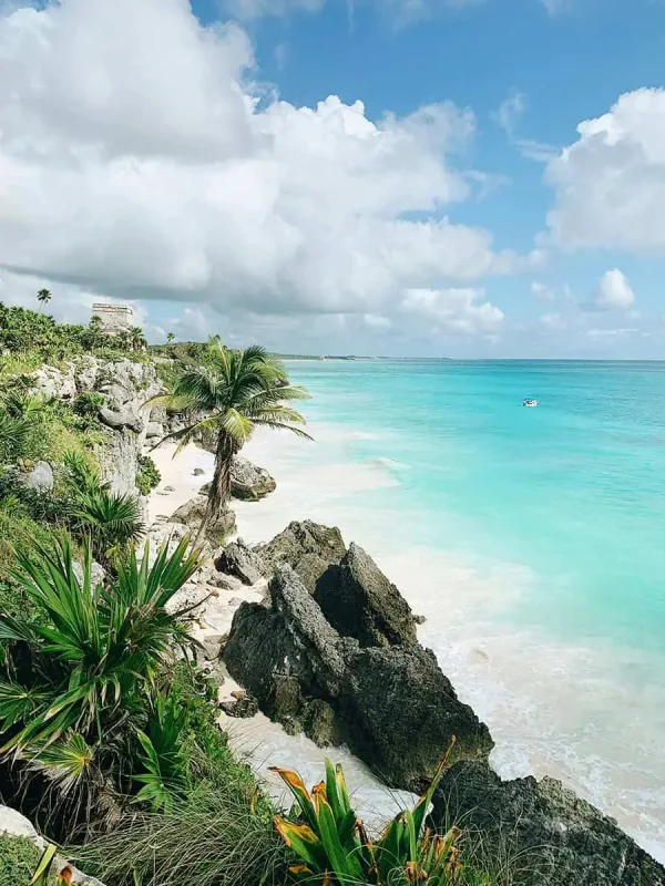 A picturesque view of Tulum's beach featuring white sandy shores and turquoise waters, framed by lush tropical vegetation and rocky formations. An ancient Mayan ruin sits atop a cliff overlooking the Caribbean Sea, under a partly cloudy sky. This image captures the unique blend of natural beauty and historical significance.