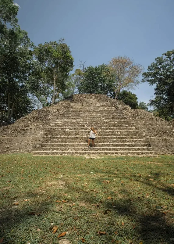tasha amy stands in front of a small but well-preserved Mayan ruin in San Ignacio. The structure, covered in vines and surrounded by lush greenery, rises against a clear blue sky.