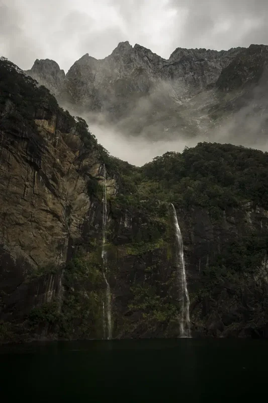 Twin waterfalls cascade down steep cliffs into dark water with mist clinging to the rocky mountains in Milford Sound, New Zealand