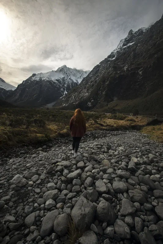 tasha amy walks across a rocky riverbed surrounded by towering snow-capped mountains in Fiordland, New Zealand
