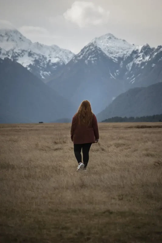 tasha amy with long hair in a red jacket walks across an open golden field toward sharp snowy peaks in eglinton valley