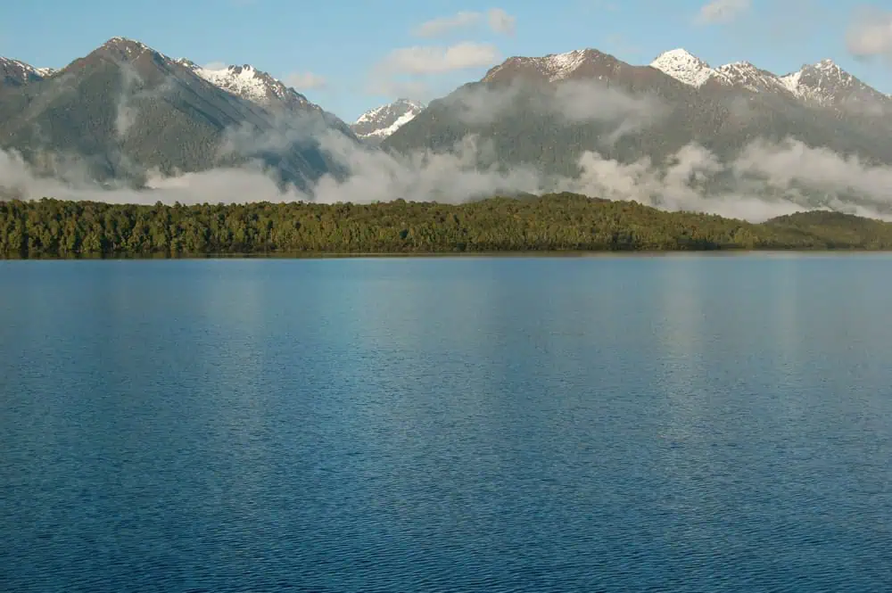 Calm waters stretch before a line of forest and mountains partly covered in mist at Manapouri Lake
