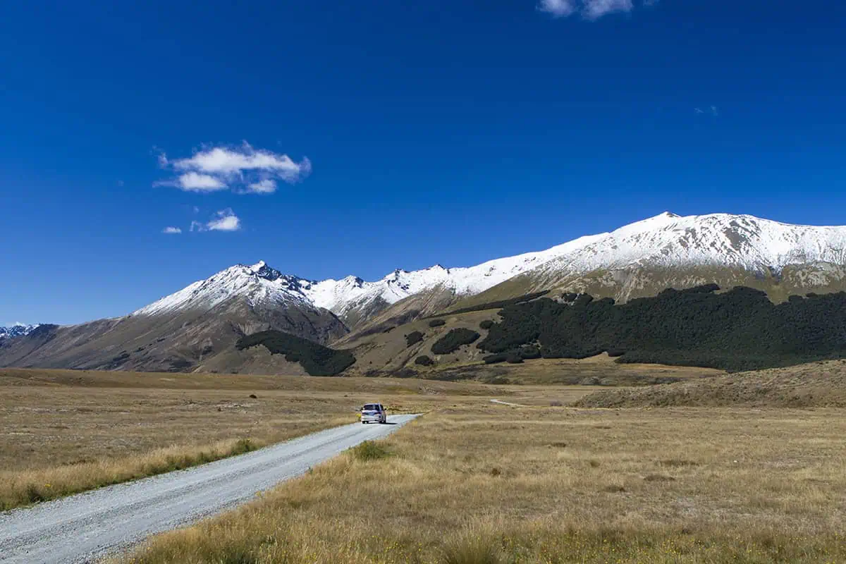 A car drives along a gravel road toward snow-capped peaks under a blue sky in New Zealand’s Lord of the Rings landscape.