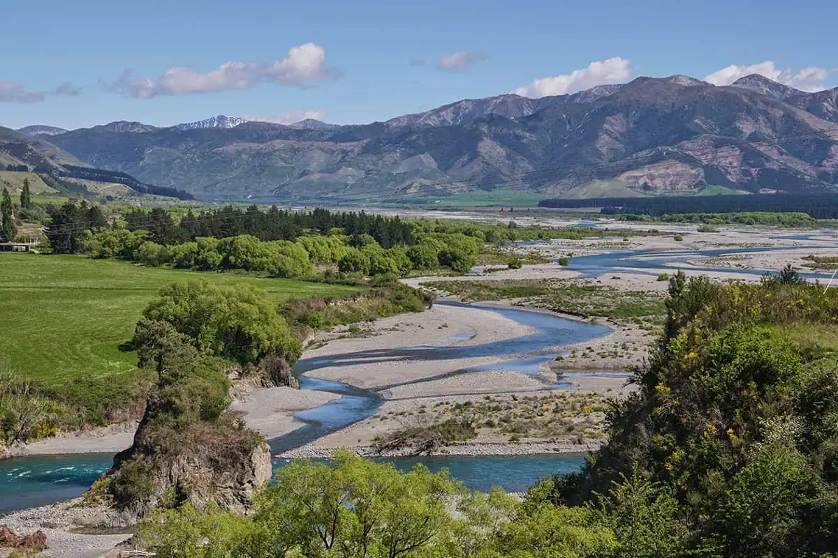 A braided Waiau River winds through fields and rocky banks with mountains rising in the background