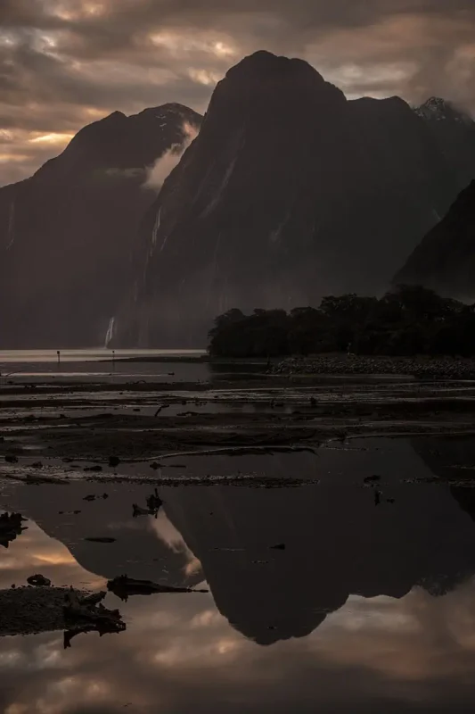 Dark shot of cliffs and peaks reflected in the shore, with waterfall streams visible under the sky.