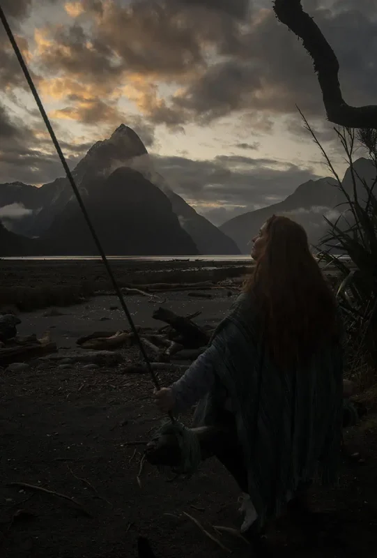 tasha amy in a poncho sits on the rustic Milford Sound swing facing Mitre Peak, the scene bathed in soft, diffused light from the overcast sky and evening glow.