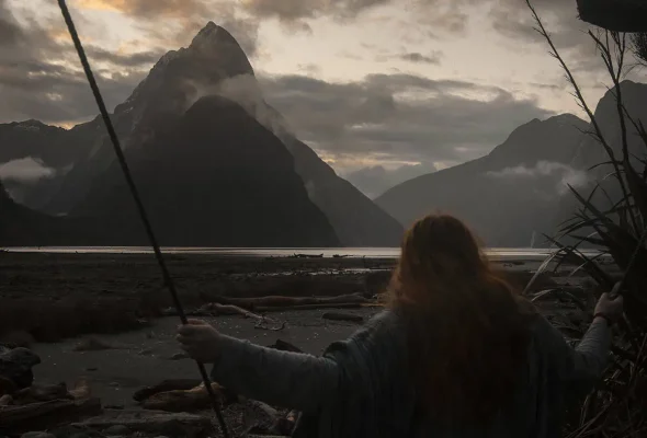 tasha amy with long red hair swings at sunset, facing the dramatic silhouette of Mitre Peak in Milford Sound with mist and low clouds rolling over the surrounding mountains.