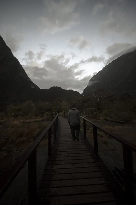 A lone man walks a wooden boardwalk framed by cliffs under a gray sky.
