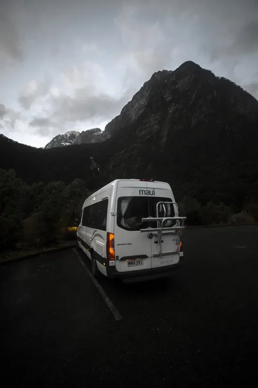 Our white Maui campervan with a bike rack is parked near the forest in the Milford Sound area, just before sunset.