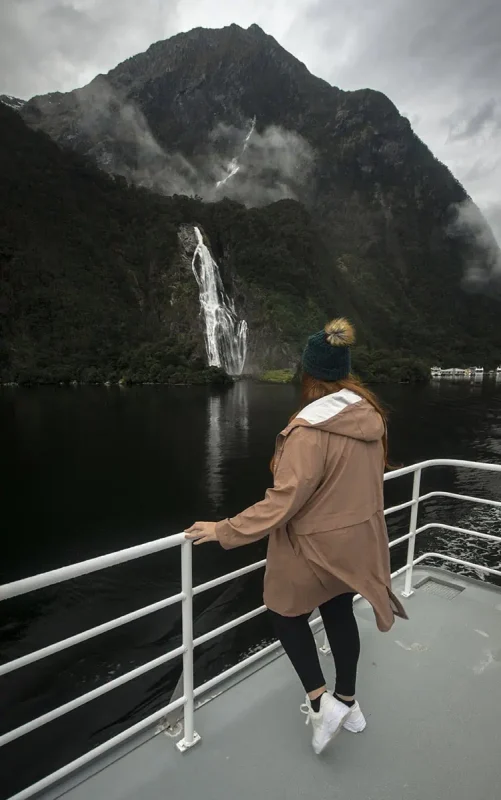 Tasha Amy, in a tan jacket and beanie, stands on a boat deck, gazing at Bowen Falls cascading from a cliff into the waters of Milford Sound.