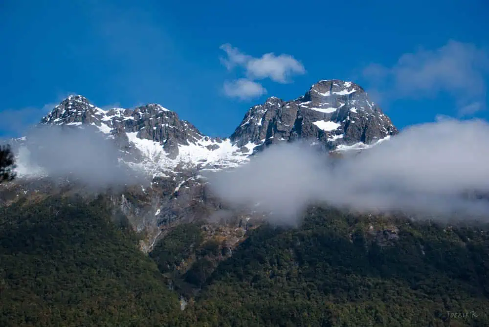 A close-up view of majestic, snow-capped Earl Mountain Peaks against a brilliant blue sky. The rugged mountain tops are partially obscured by low-hanging clouds, enhancing the dramatic contrast between the pristine white snow and the dark, rocky surfaces. The lush, green foothills below add a touch of softness to the otherwise stark landscape.