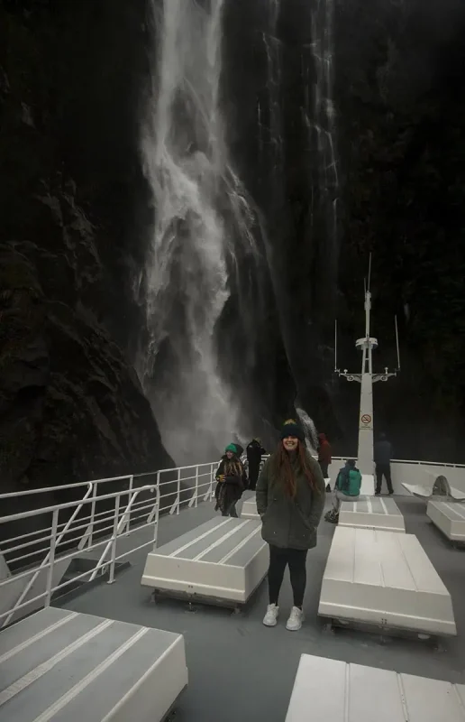A group of people on a boat deck near a powerful waterfall in Milford Sound. The waterfall cascades down a steep, rocky cliff, creating a misty spray that envelops the area. The passengers, dressed warmly, seem captivated by the natural spectacle.