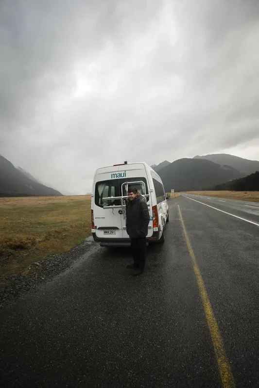 A person standing next to a white "Maui" campervan on a wet, overcast day. The campervan is parked on a roadside with a vast, open field and misty mountains in the background. The gray sky and damp ground create a moody, traveler's scene.