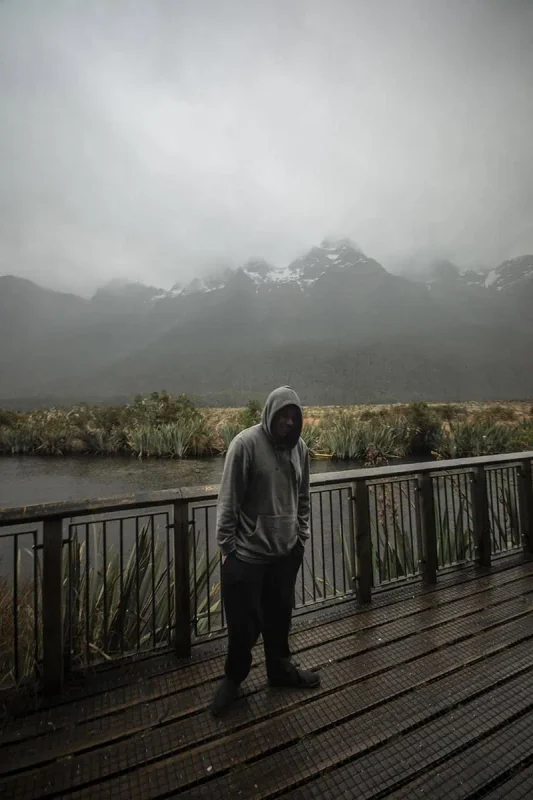 A person in a gray hoodie stands on a wooden platform overlooking Mirror Lakes in Milford Sound. The platform is surrounded by dense greenery and offers a view of the misty, snow-capped mountains in the background. The gray, cloudy sky and the reflective surface of the lake create a serene and contemplative atmosphere.