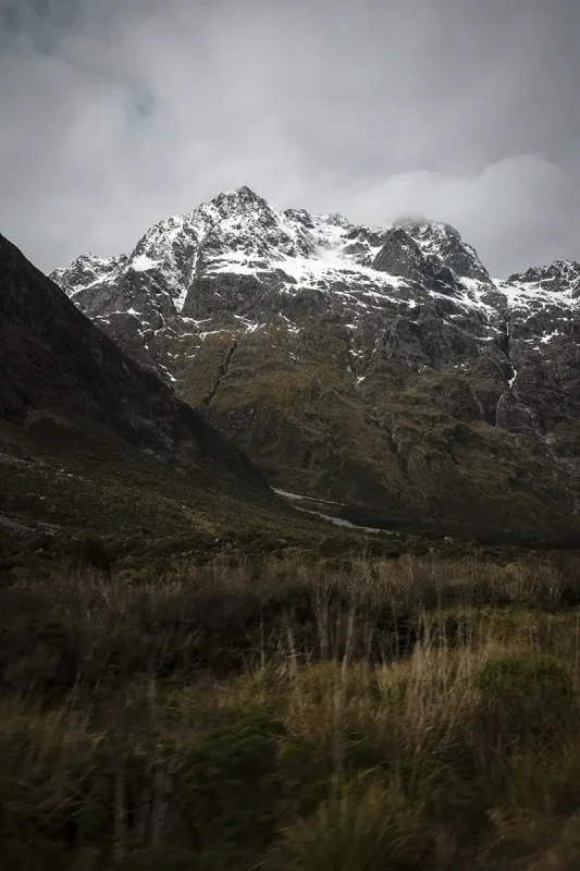 A striking view of the mountainous terrain at Monkey Creek, New Zealand. The rocky slopes are streaked with patches of snow, contrasting against the green and brown hues of the vegetation below. The cloudy sky overhead adds a moody and atmospheric quality to the scene, emphasizing the raw beauty of the rugged landscape.