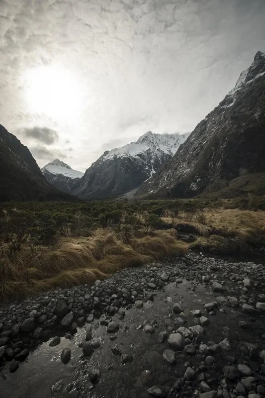 A small stream flows over a rocky bed in the foreground at Monkey Creek, New Zealand, leading the eye towards a backdrop of towering, snow-covered mountains. The landscape is a mix of golden grasses and green shrubs, contrasting with the dark, imposing peaks under a cloudy sky. The scene is serene and captivating, highlighting the natural beauty of the area.