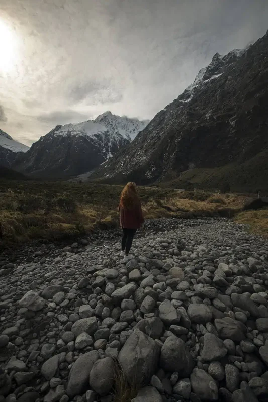 tasha amy with long, red hair walks along a rocky streambed at Monkey Creek, New Zealand. She is dressed in a rust-colored sweater and black pants, and the rugged mountains rise dramatically behind her. The overcast sky and misty atmosphere enhance the wild, untamed beauty of the landscape, emphasizing the grandeur of the natural setting.