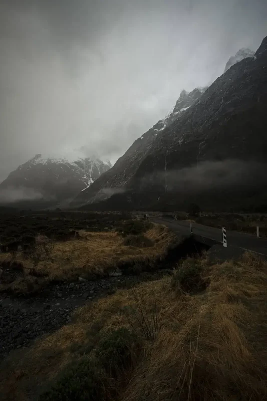 A winding road cuts through the rugged landscape of Monkey Creek, New Zealand, bordered by golden grasses and green shrubs. The road leads towards mist-shrouded mountains that loom overhead, their rocky surfaces streaked with white snow. The cloudy sky adds to the dramatic and remote feel of the scene.