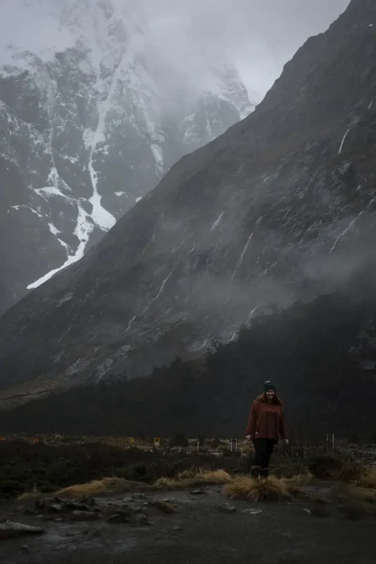 tasha amy dressed in a rust-colored sweater, black pants, and a beanie stands in the foreground of Monkey Creek. She is surrounded by rugged terrain and mist-covered mountains that rise sharply behind her. The dark, overcast sky and low clouds enhance the wild and untamed beauty of the landscape.
