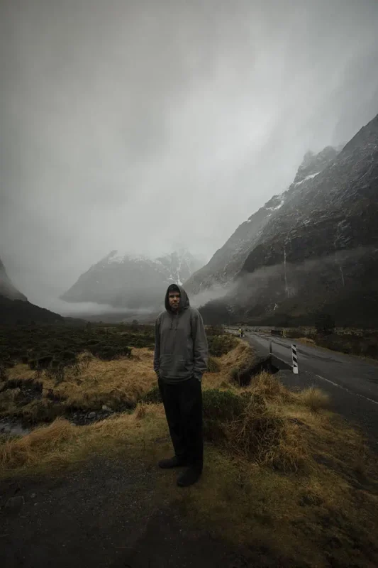 A man in a grey hoodie stands on a grassy, rocky path at Monkey Creek, New Zealand. Behind him, a winding road disappears into the misty, mountainous background. The dark, imposing mountains are partially obscured by low-hanging clouds, creating a mysterious and atmospheric scene.