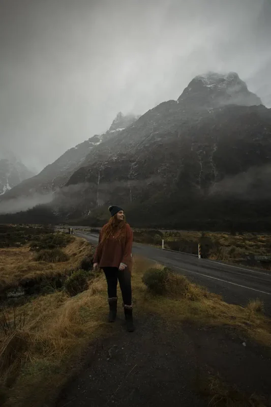tasha amy wearing a rust-colored sweater and black pants stands near a winding road in Monkey Creek. The road leads towards dramatic, mist-covered mountains that tower in the background. The cloudy sky and rugged landscape create a sense of solitude and natural beauty.