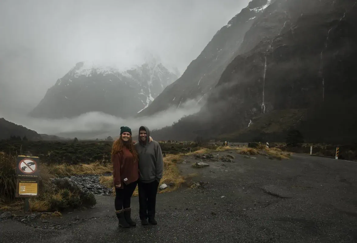 A couple poses together at Monkey Creek, New Zealand, with the woman wearing a rust-colored sweater and beanie and the man in a grey hoodie. They stand in front of a dramatic landscape of mist-covered mountains and a lush valley. A sign in the foreground advises against feeding the local kea birds, adding a touch of local wildlife awareness to the scene.