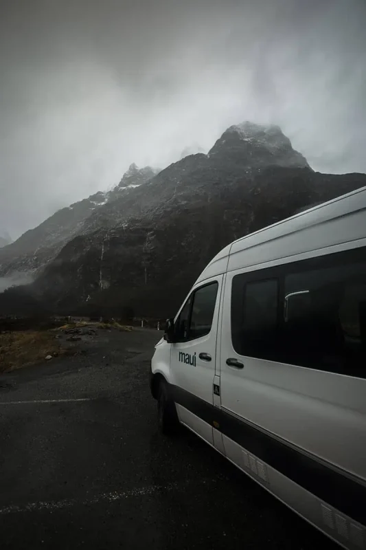 A close-up view of a white "Maui" camper van parked up on the side of the road to milford sound. The van is set against the backdrop of towering, mist-covered mountains. The dark, rocky peaks and overcast sky create a striking contrast with the bright, white van, highlighting the ruggedness of the natural environment.