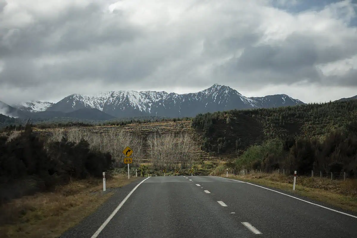 An open road leading towards snow-dusted mountain ranges under a cloudy sky. The sign on the side of the road indicates a sharp turn.