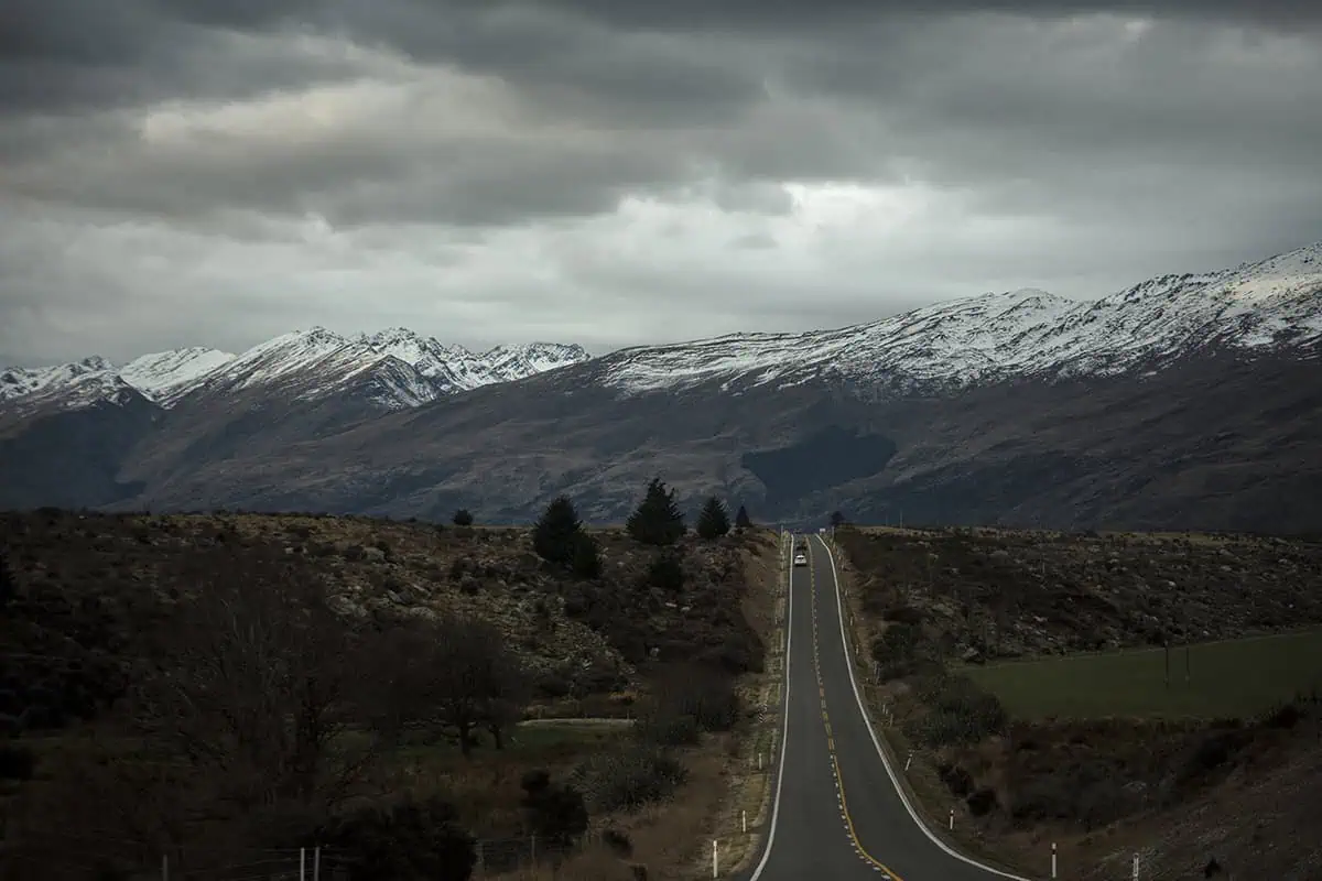 An open road stretches towards the distant, snow-capped mountains under a cloudy sky, with barren, rugged terrain on either side. This scene captures the remote and scenic drive from Queenstown to Milford Sound.