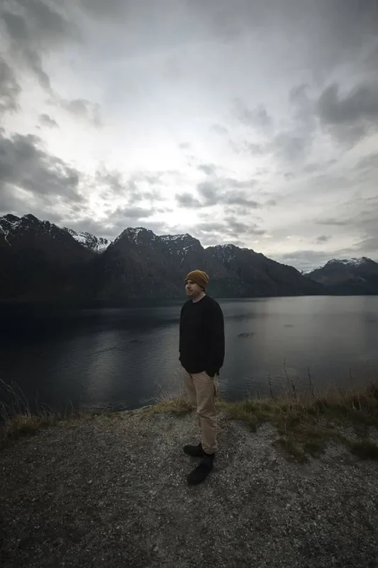 A person stands by the edge of a lake, with snow-capped mountains in the background under a cloudy sky. The reflective lake and towering peaks emphasize the tranquil yet majestic landscape encountered on the journey