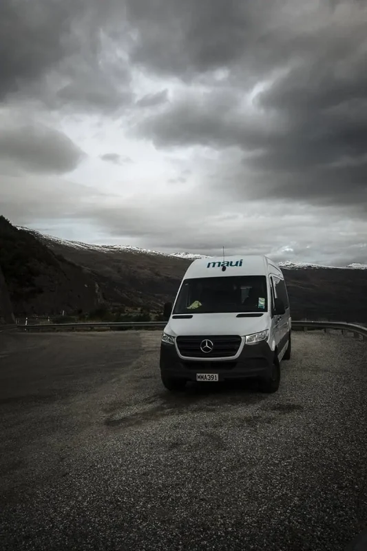 A white camper van marked "maui" is parked on a gravel pull-off area, with dramatic, overcast skies and mountain views in the background. The camper van represents the adventurous spirit of road-tripping from Queenstown to Milford Sound.
