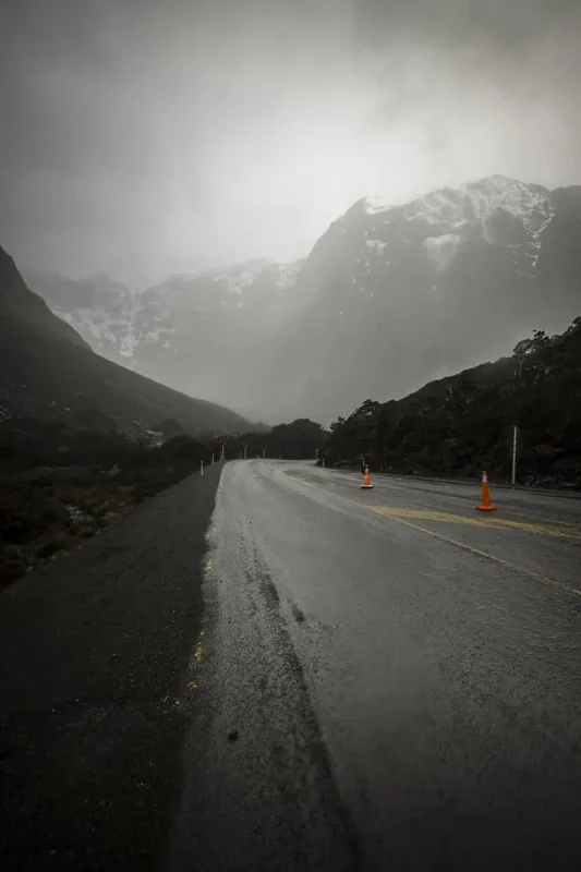 A rainy, winding mountain road flanked by tall, rugged peaks and traffic cones marking the path. The mist and rain emphasize the dramatic weather conditions on the route from Queenstown to Milford Sound.