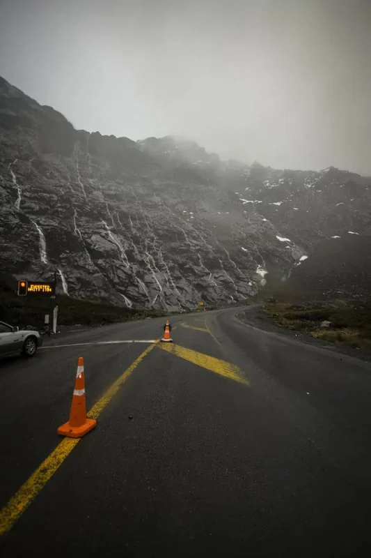 A closer view of the winding mountain road, showing the steep rock face on one side and the misty atmosphere. Traffic cones and a sign with blurred text indicate the road conditions