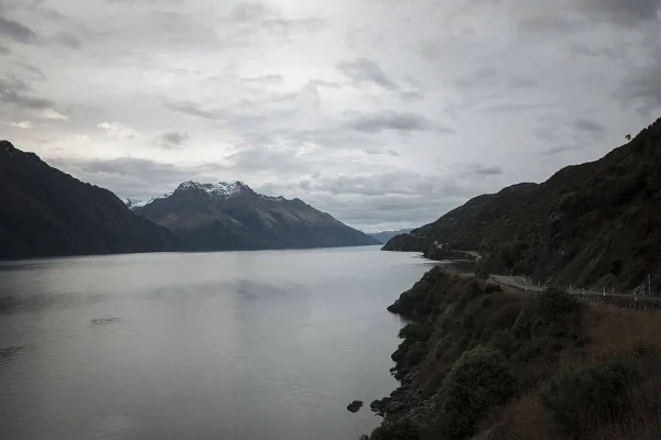A serene view of Lake Wakatipu, with calm waters reflecting the overcast sky and snow-capped mountains in the background. The winding road along the lake hints at the journey from Queenstown to Milford Sound.