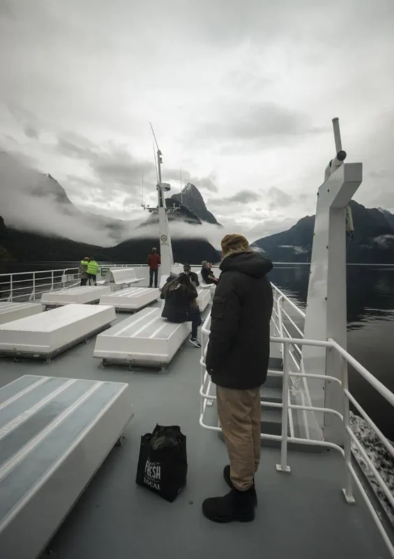 People stand on the deck of a boat, taking in the majestic views of Milford Sound's steep cliffs and cloudy sky.