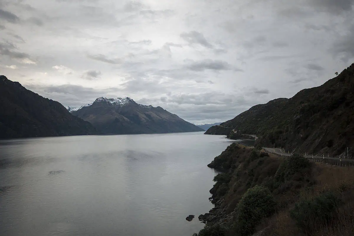 A serene view of Lake Wakatipu, with calm waters reflecting the overcast sky and snow-capped mountains in the background. The winding road along the lake hints at the journey from Queenstown to Milford Sound.