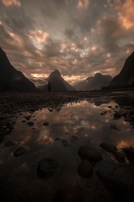 reflective pool with Milford Sound's iconic Mitre Peak in the background at sunset. The sky is ablaze with orange and pink hues, reflected in the still waters, creating a serene yet dramatic scene.