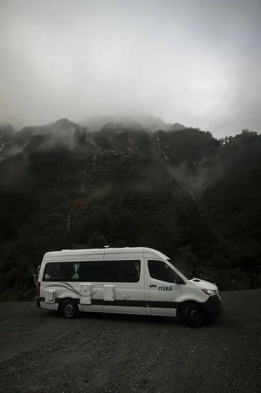 A white camper van is parked against the backdrop of a misty, mountainous landscape in Milford Sound. The mountain is partially obscured by low clouds, with thin waterfalls trickling down its slopes, creating a picturesque yet typical rainy day in Milford Sound.