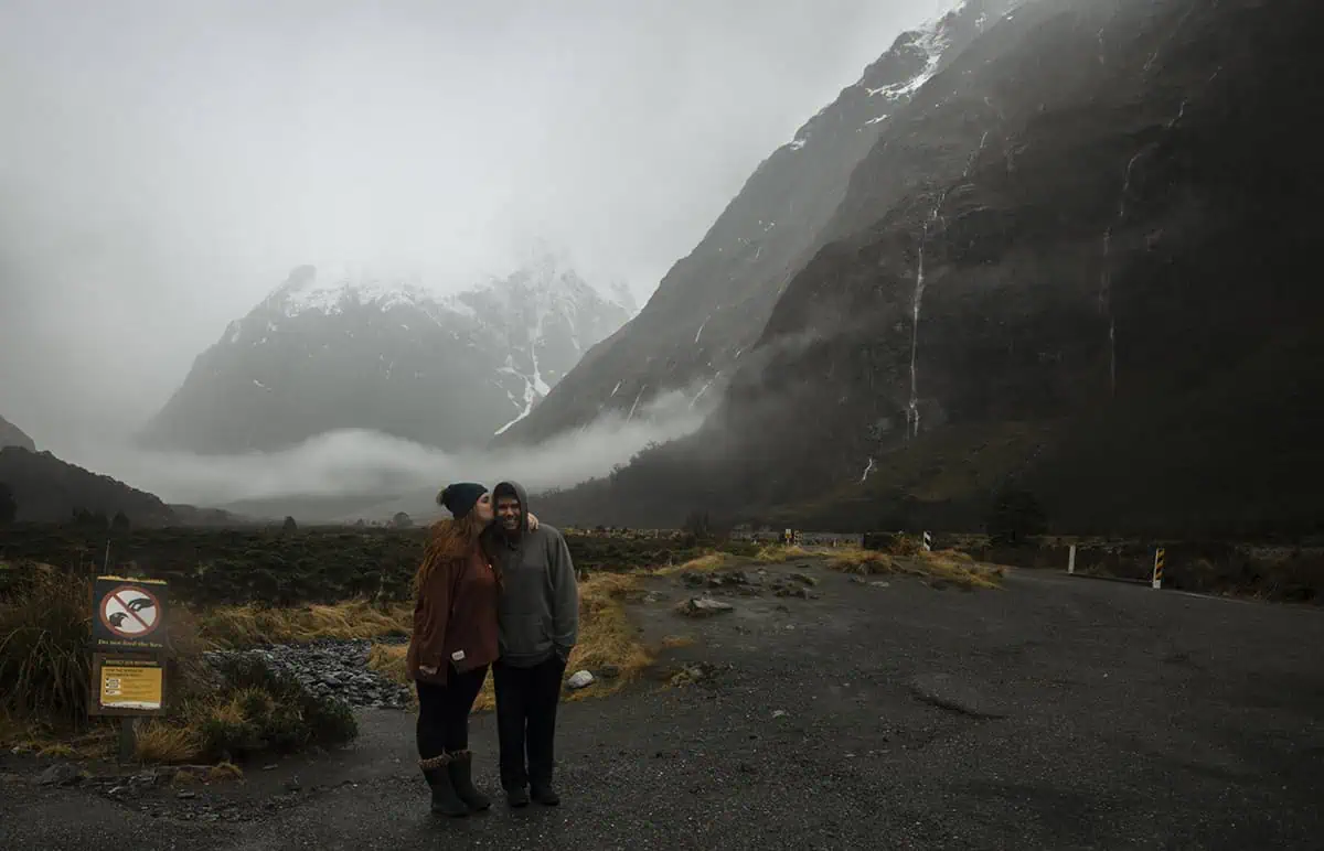 A couple stands close together, with one kissing the other's cheek, against a backdrop of misty mountains in Milford Sound. The ground is wet, and the low clouds and overcast sky highlight the rainy weather in Milford Sound.