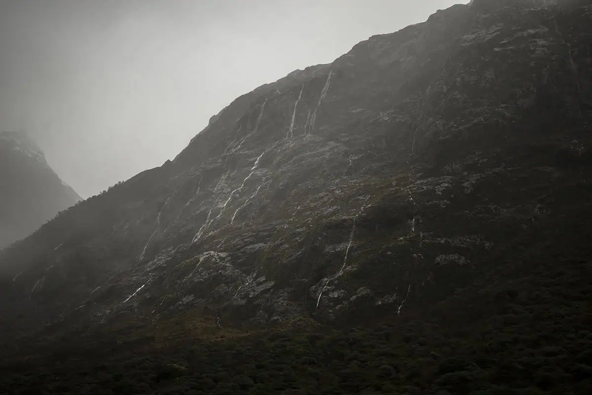 Misty mountain slopes with thin waterfalls trickling down the rugged surface in Milford Sound. The overcast sky and mist create a dramatic and atmospheric scene, typical of the rainy weather in Milford Sound.