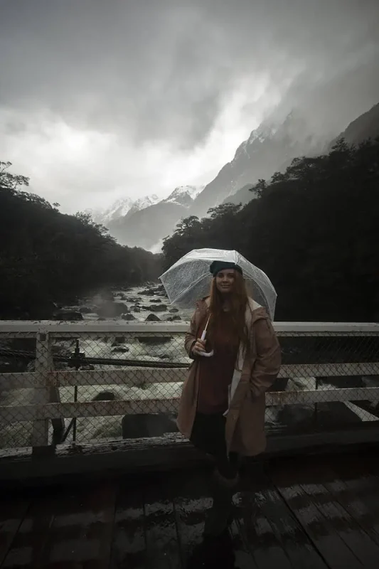 tasha amy stands on a bridge holding an umbrella, overlooking a fast-flowing river surrounded by misty mountains in Milford Sound. The overcast sky and low clouds highlight the typical rainy weather in the region.
