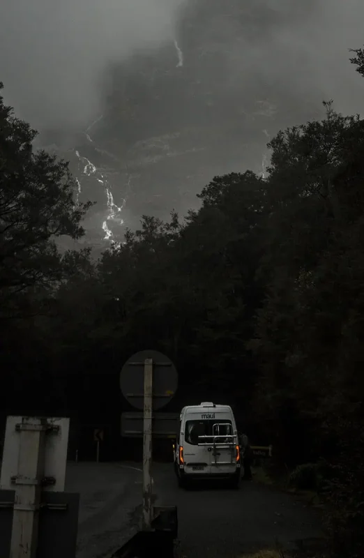 A white camper van is parked at the edge of a forest with a misty mountain backdrop in Milford Sound. The scene is enveloped in low clouds and light rain, creating a serene and atmospheric setting.