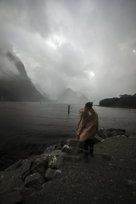 tasha amy tands on a rocky shore, looking out over the calm waters of Milford Sound, with misty mountains in the background. The scene is enveloped in a light rain, enhancing the mystical and tranquil ambiance of the fjord.