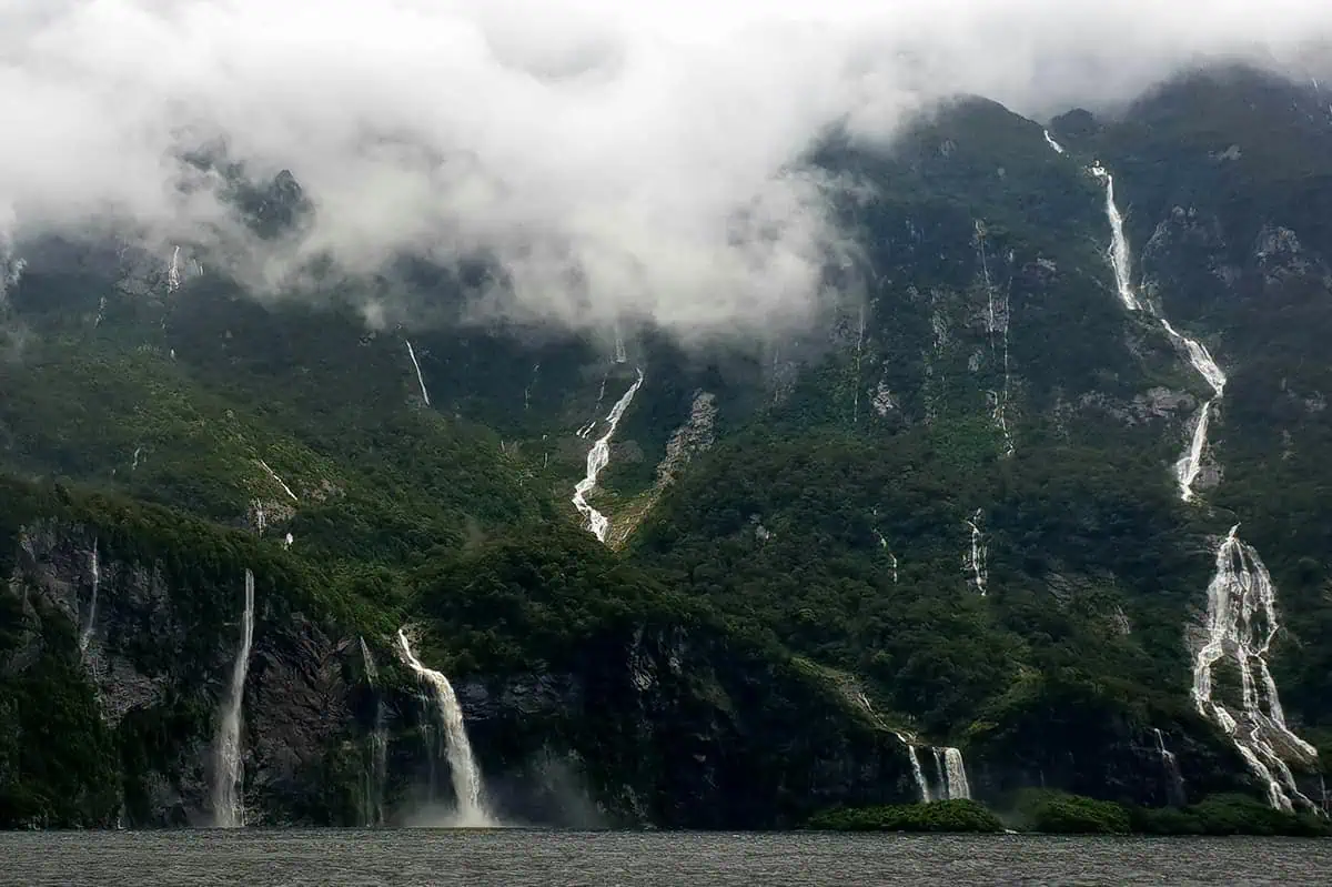 aterfalls cascade down lush, green mountains shrouded in mist and low-hanging clouds in Milford Sound. The dramatic scenery captures the raw beauty of the landscape, with multiple waterfalls visible against the dense foliage. This image perfectly showcases the ethereal atmosphere of rain in Milford Sound.