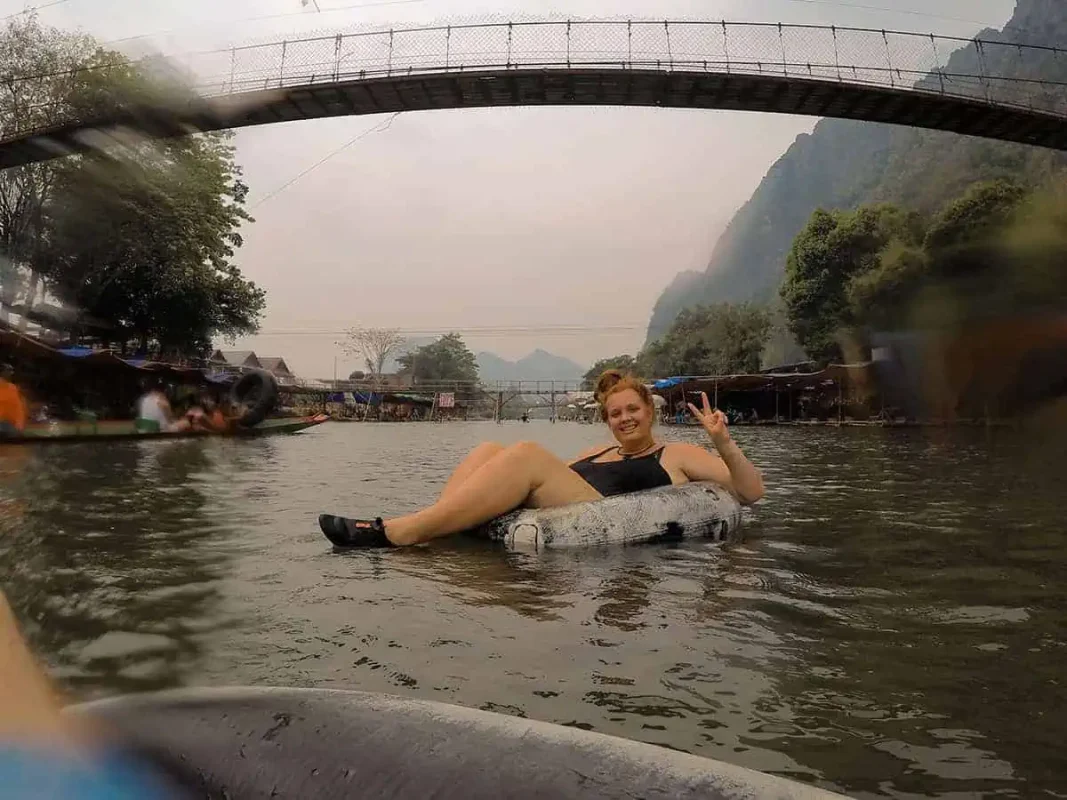 Tasha Amy smiling and making a peace sign while floating on a tube in a river under a bridge with a rustic village backdrop in Laos