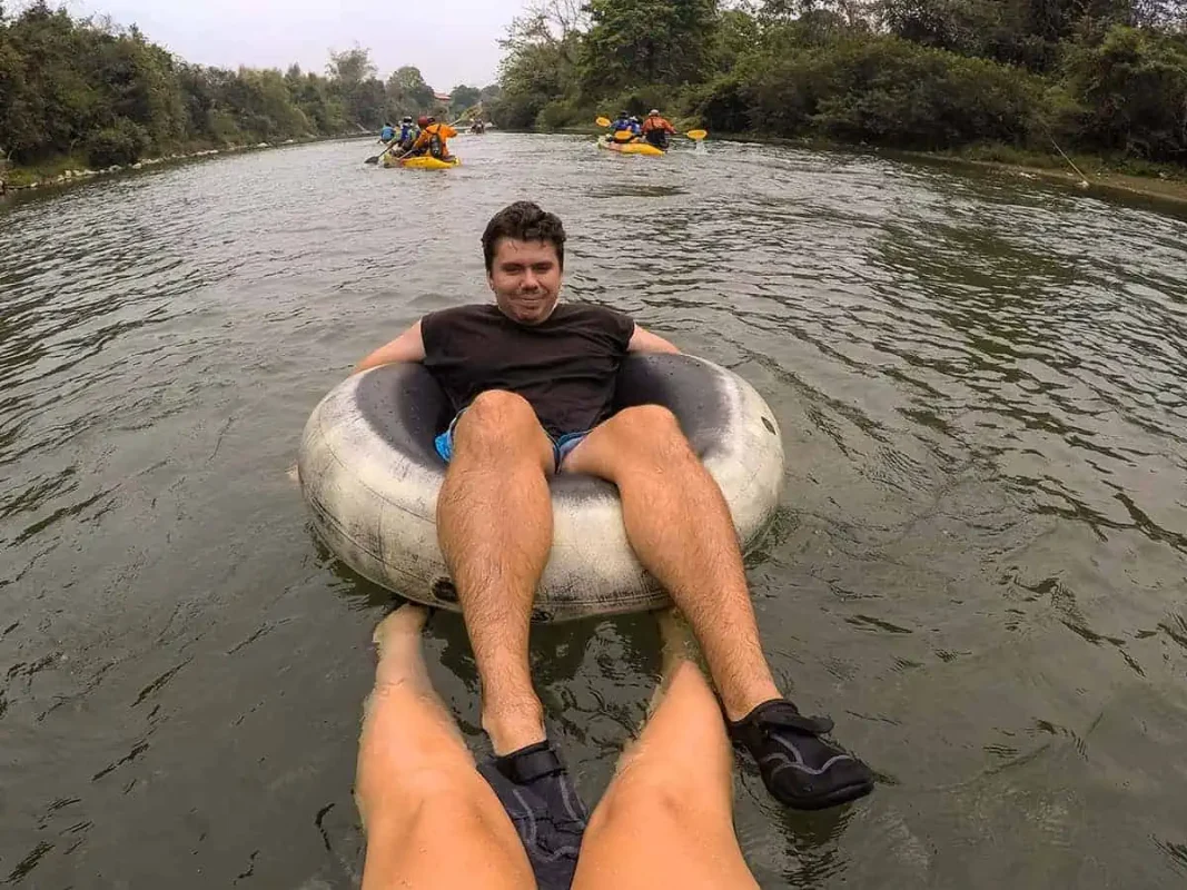 A man lounging in a tube on a river with other people kayaking in the background, surrounded by dense foliage in Vang Vieng.
