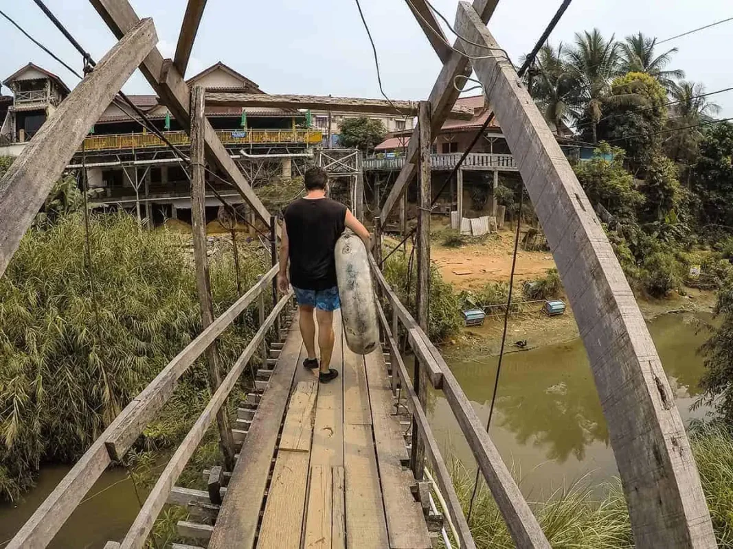 A person walking on a wooden bridge carrying a tube, with traditional houses on stilts and dense vegetation on either side.