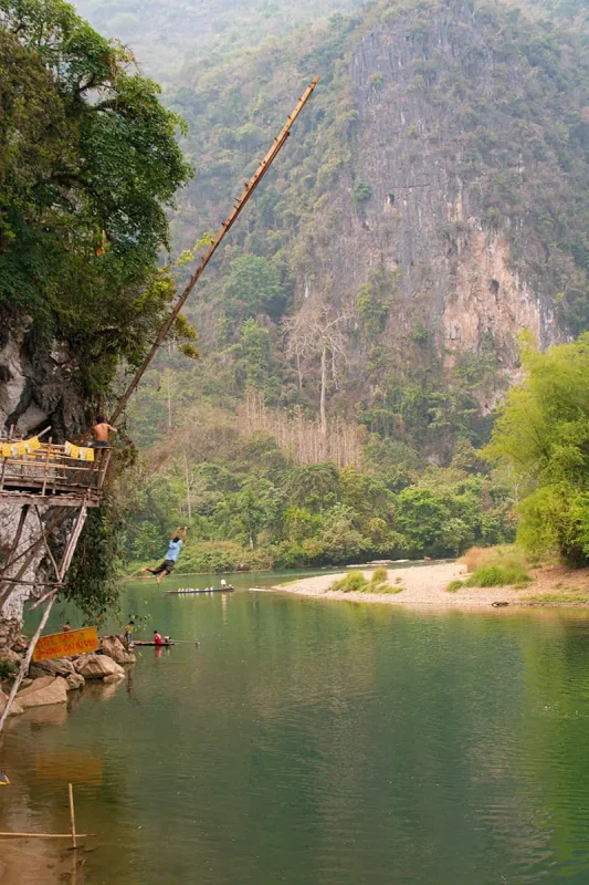 A daring individual swings from a high platform on a rope swing above a tranquil river surrounded by steep cliffs and greenery in Laos.